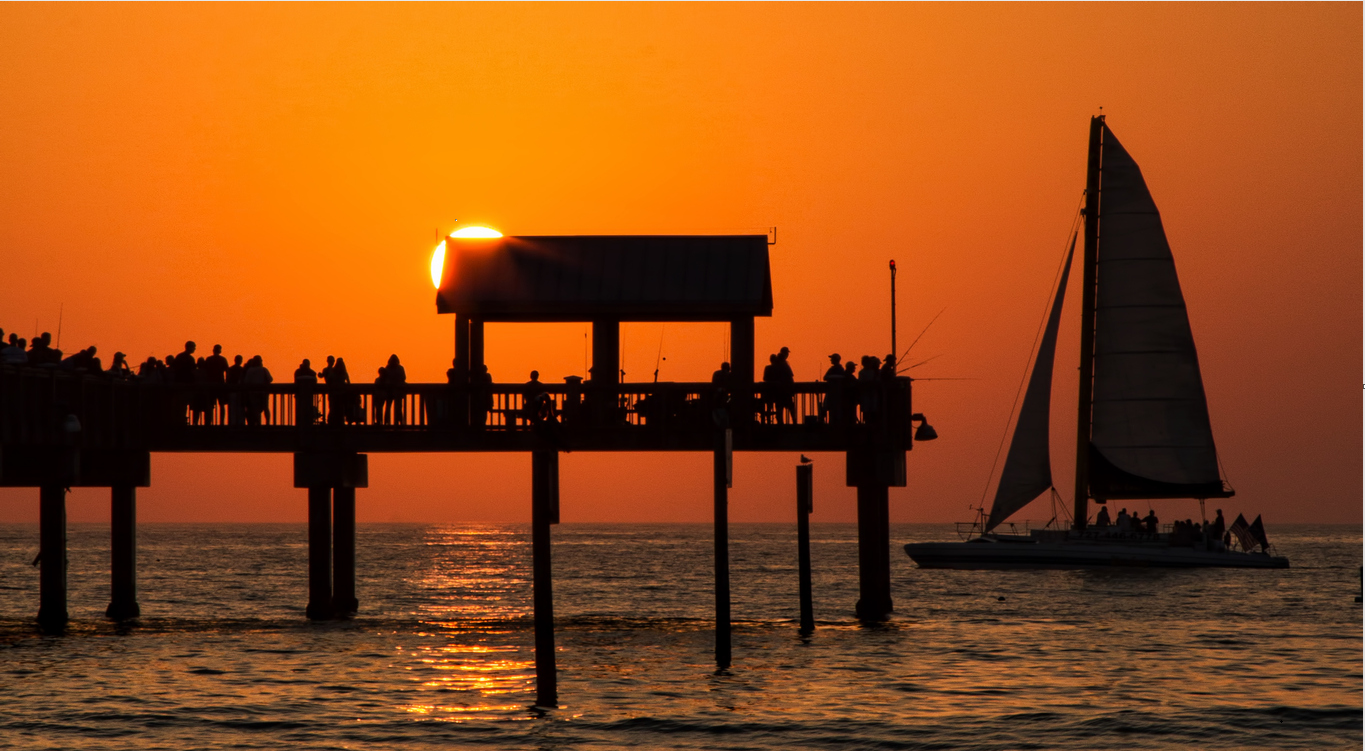 Sunset over the ocean appears behind a dock with a sailboat on the horizon.