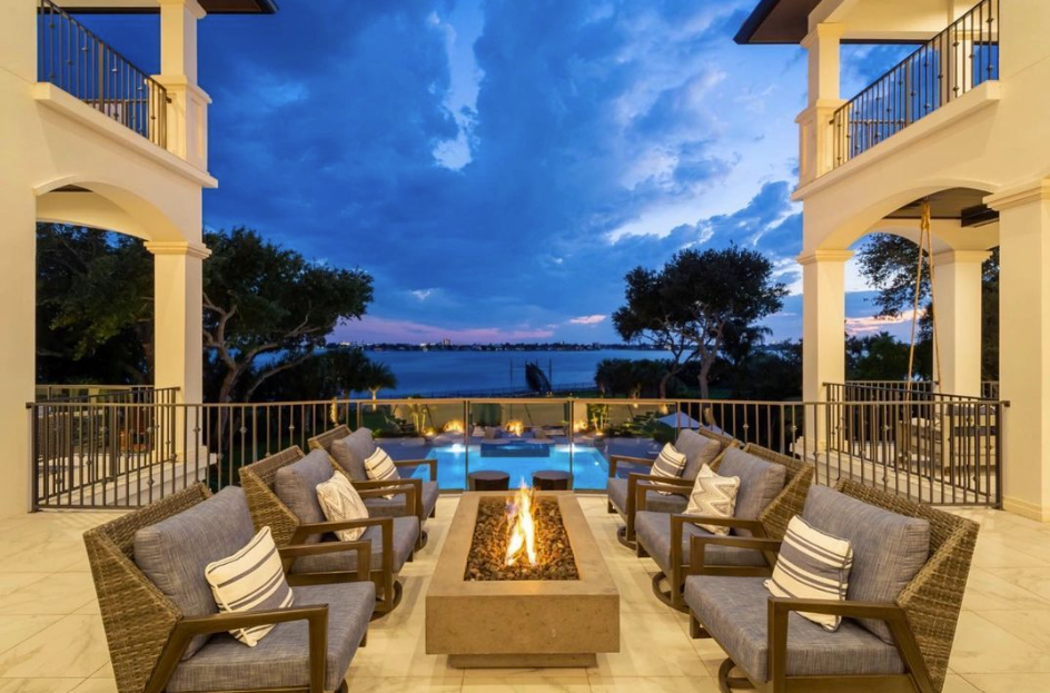 An outdoor fireplace with comfortable seating overlooks a blue swimming pool and water view